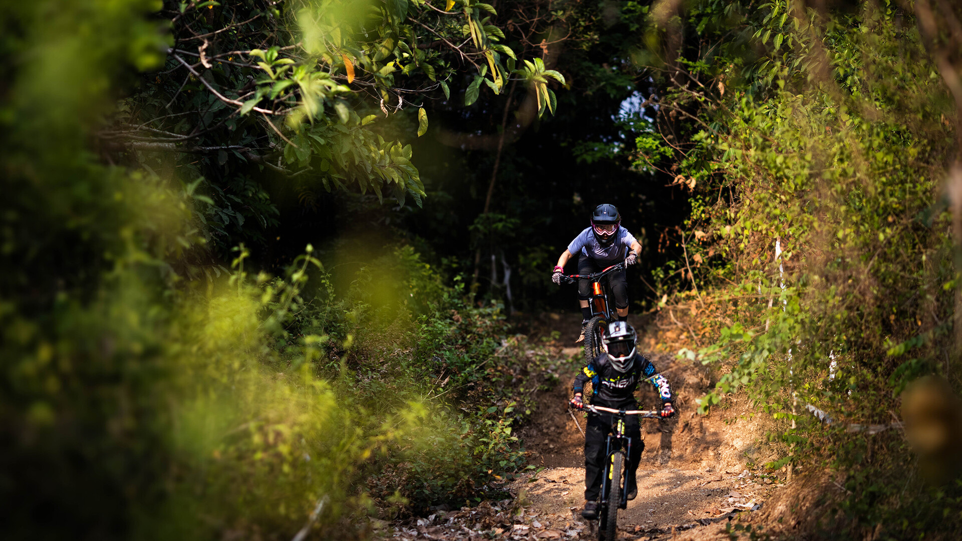 Two mountain bikers riding down a narrow, tree-lined trail. The front rider is wearing black gear with a helmet, while the rider behind is dressed in grey with goggles.